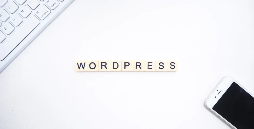 Fichas de letras que forman la palabra WordPress sobre una mesa blanca, junto a un teclado y un móvil